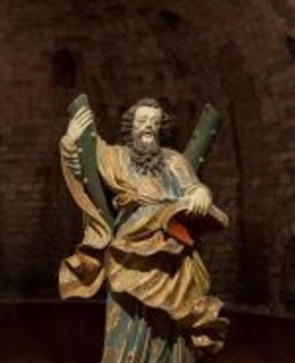 Estátua de figura religiosa segurando dois bastões e um livro, em ambiente escuro.