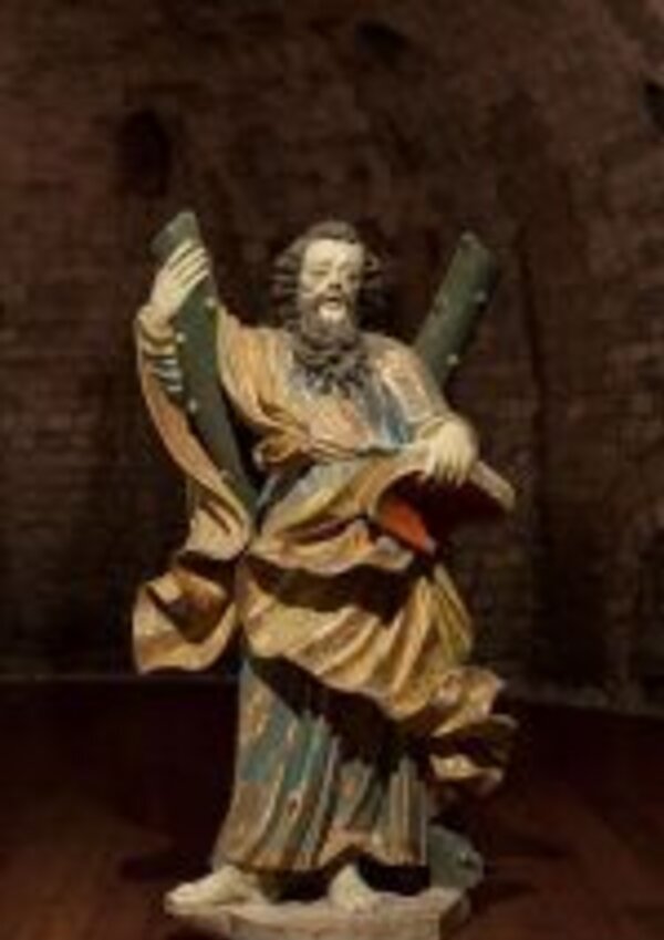 Estátua de figura religiosa segurando dois bastões e um livro, em ambiente escuro.