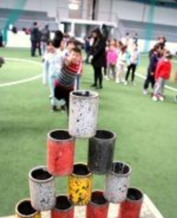 Piramide de latas coloridas em campo, crianças brincando ao fundo.
