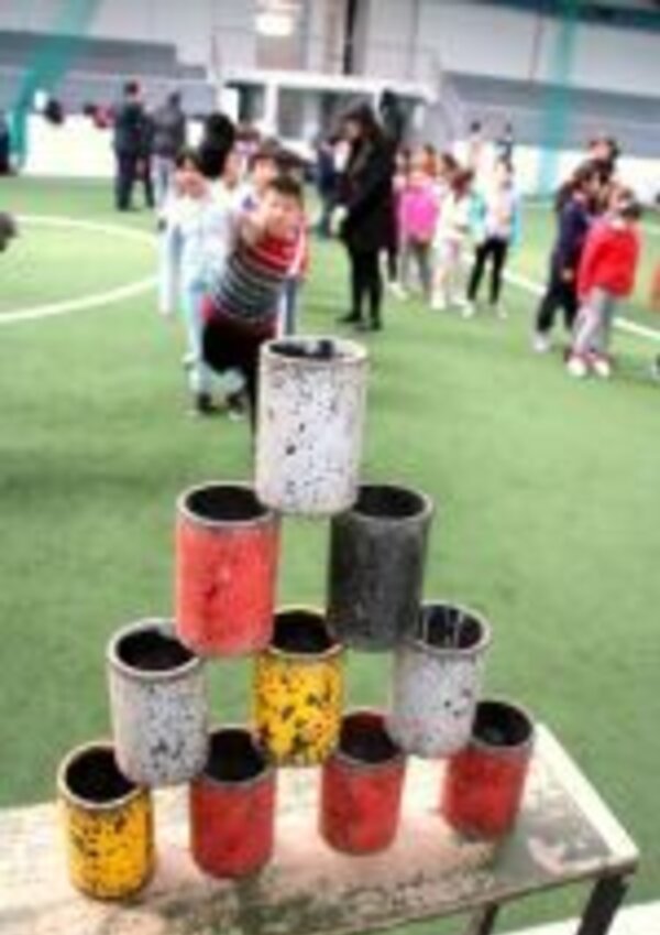 Piramide de latas coloridas em campo, crianças brincando ao fundo.