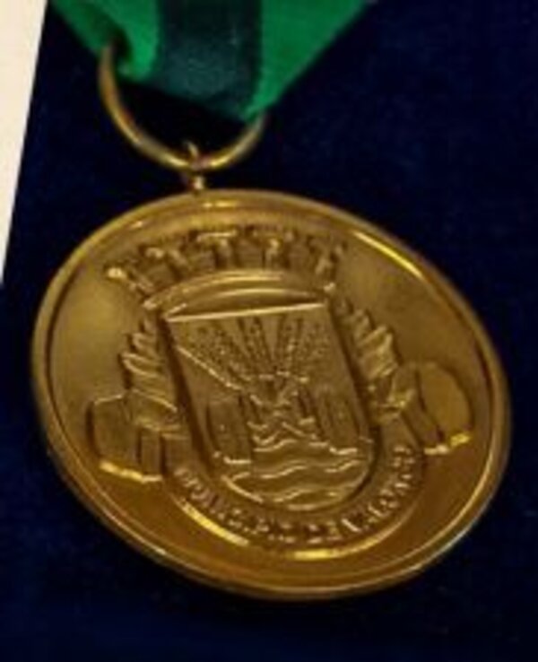 Medalha dourada com um brasão e fita verde, sobre fundo azul.