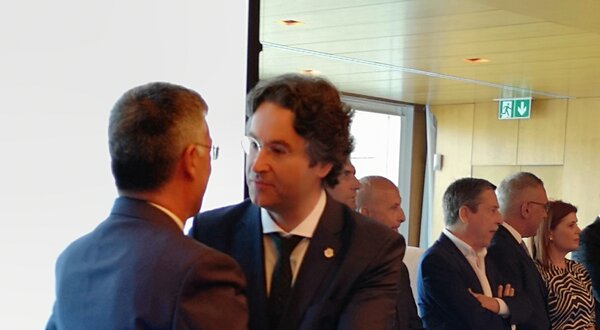 Dois homens em traje formal a cumprimentar-se num evento, com outros ao fundo.