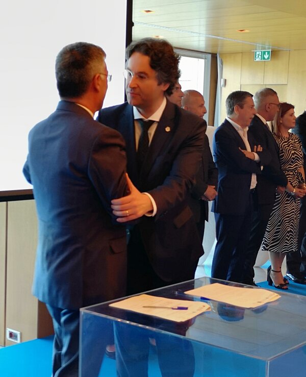 Dois homens em traje formal a cumprimentar-se num evento, com outros ao fundo.