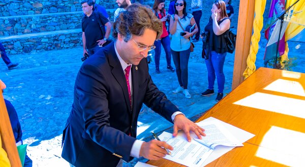 Homem a assinar documentos numa mesa, com pessoas ao fundo.