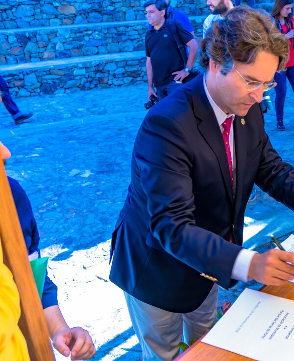 Homem a assinar documentos numa mesa, com pessoas ao fundo.