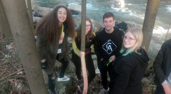 Grupo de jovens a explorar a natureza junto a um rio, com árvores ao redor.