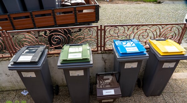 Quatro contentores de lixo coloridos para reciclagem, alinhados numa calçada.
