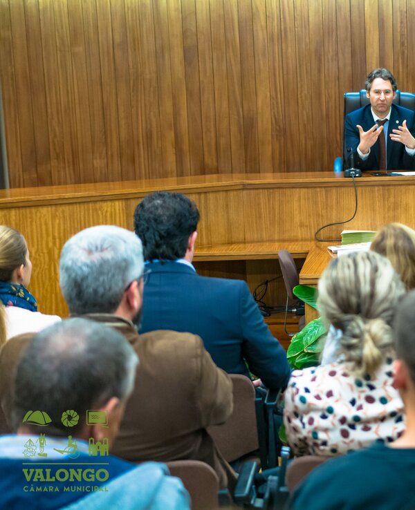 Reunião em câmara municipal com várias pessoas a ouvir um orador.