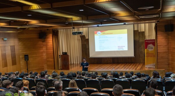 Palestra em auditório com público atento e apresentador ao centro.