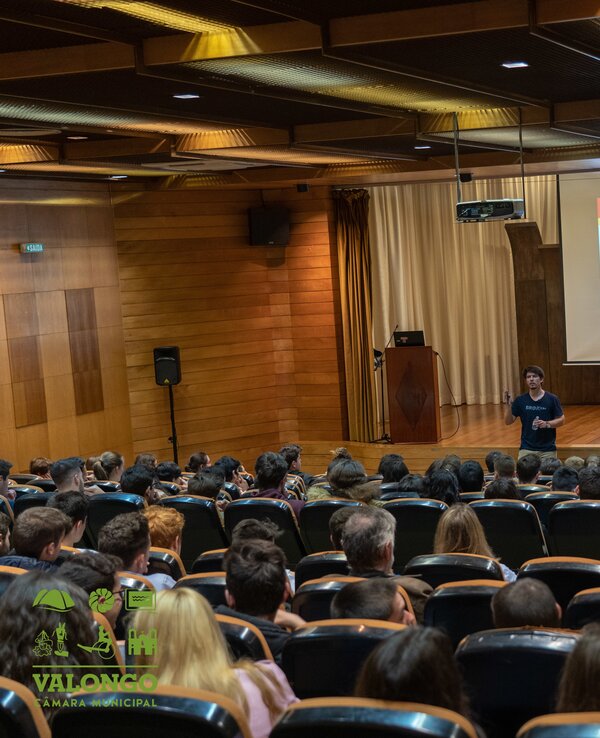 Palestra em auditório com público atento e apresentador ao centro.