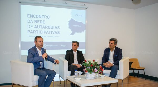 Três homens sentados em sofás, discutindo num evento sobre autarquias participativas.