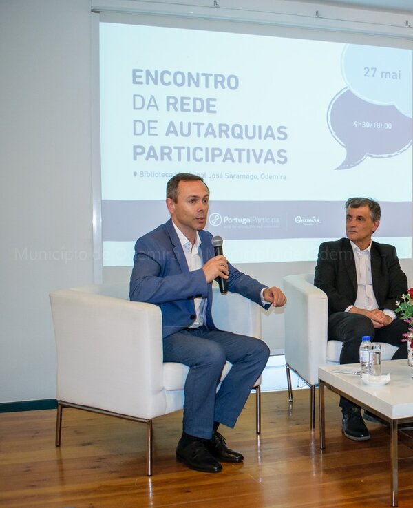 Três homens sentados em sofás, discutindo num evento sobre autarquias participativas.