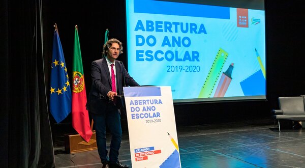 Um orador em palco com bandeiras ao fundo, apresentando a abertura do ano escolar.