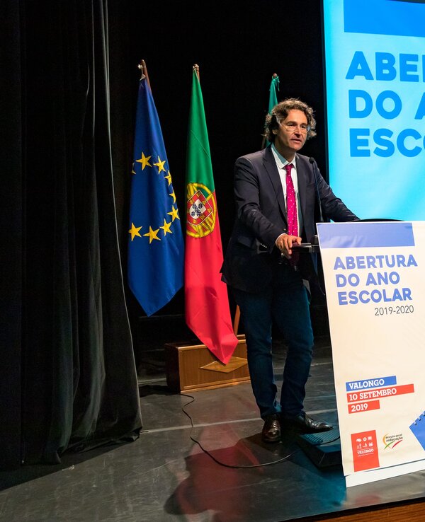 Um orador em palco com bandeiras ao fundo, apresentando a abertura do ano escolar.