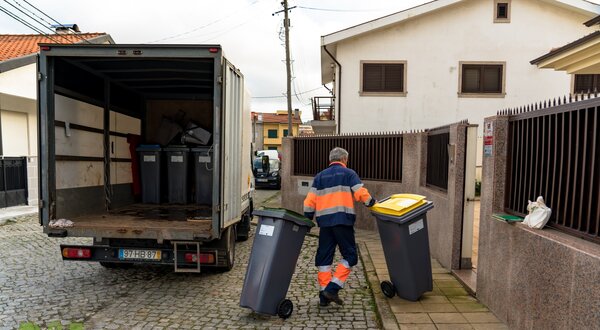 Um trabalhador a transportar contentores de lixo numa rua residencial.