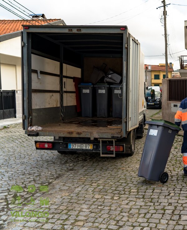 Um trabalhador a transportar contentores de lixo numa rua residencial.