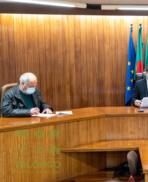 Três homens sentados à volta de uma mesa em reunião, com bandeiras ao fundo.