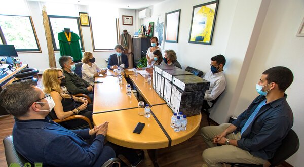 Reunião em mesa oval com várias pessoas, documentos e garrafas de água.