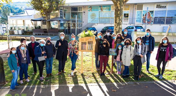 Grupo de pessoas sorridentes em um parque, usando máscaras, ao lado de uma estante.