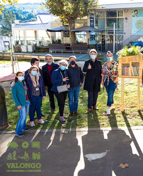 Grupo de pessoas sorridentes em um parque, usando máscaras, ao lado de uma estante.