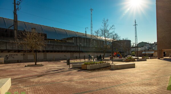 Praça ampla com pavimento de ladrilhos, árvores e pessoas a passear sob o sol.
