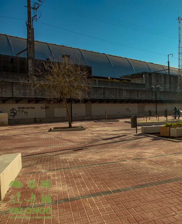 Praça ampla com pavimento de ladrilhos, árvores e pessoas a passear sob o sol.
