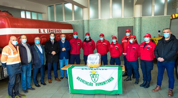 Grupo de bombeiros e autoridades em frente a um veículo, com bandeira da associação.