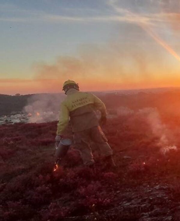Bombeiro a combater um incêndio ao pôr do sol, com fumo e chamas visíveis.