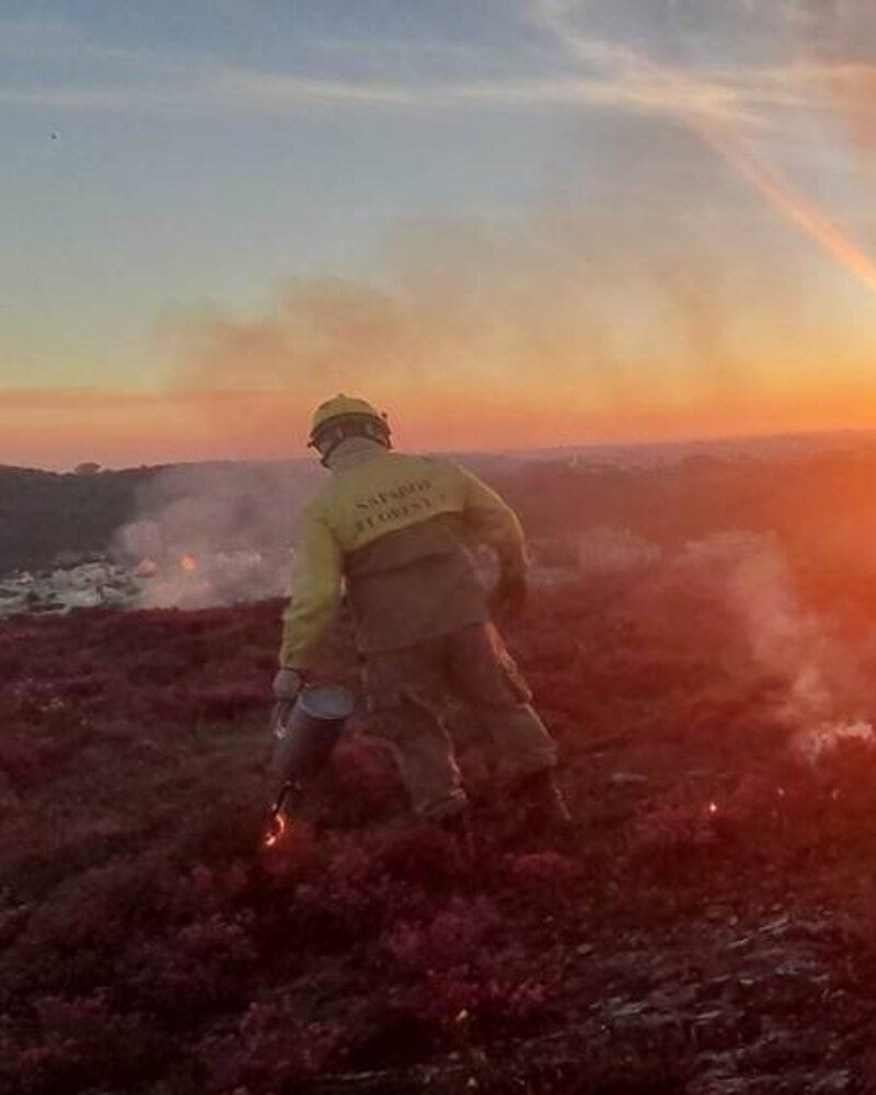 Bombeiro a combater um incêndio ao pôr do sol, com fumo e chamas visíveis.