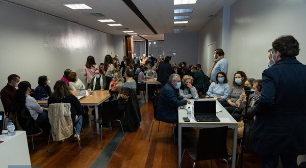Sala cheia de pessoas sentadas e em pé, a conversar e a trabalhar em grupo.