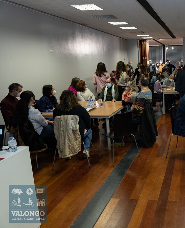 Sala cheia de pessoas sentadas e em pé, a conversar e a trabalhar em grupo.