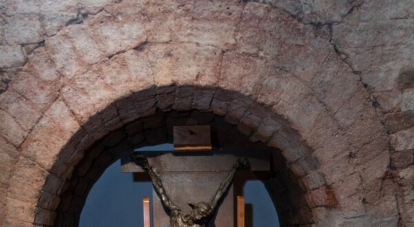 Escultura de bronze de uma figura crucificada, exposta em um arco de pedra.