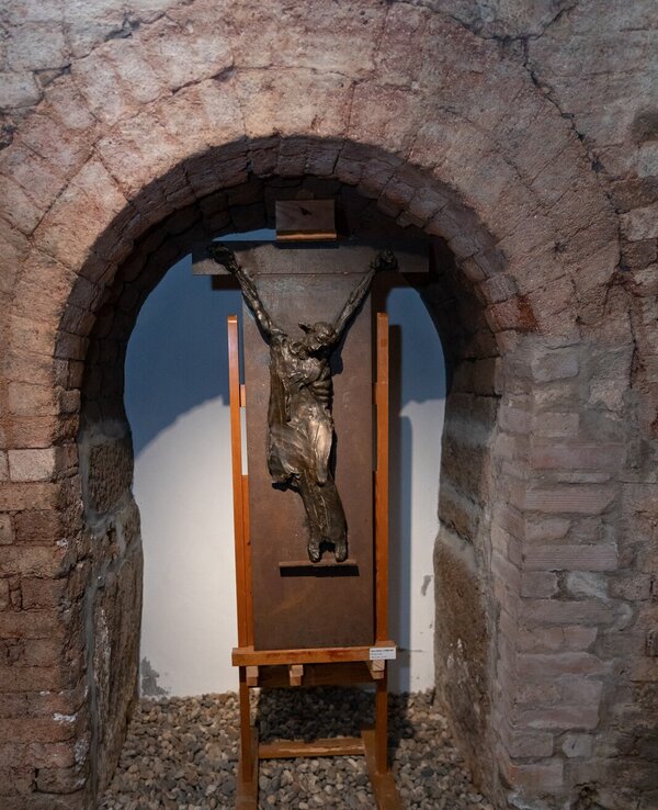 Escultura de bronze de uma figura crucificada, exposta em um arco de pedra.