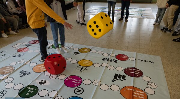 Jogadores lançam dados grandes sobre um tabuleiro colorido no chão.