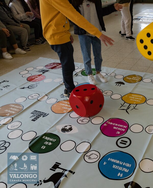 Jogadores lançam dados grandes sobre um tabuleiro colorido no chão.