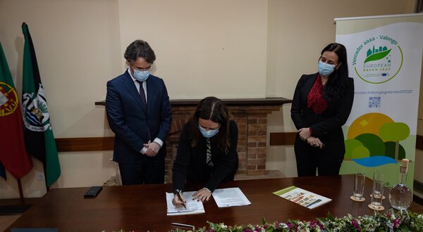 Uma mulher assina documentos em uma cerimónia, com dois homens ao fundo.