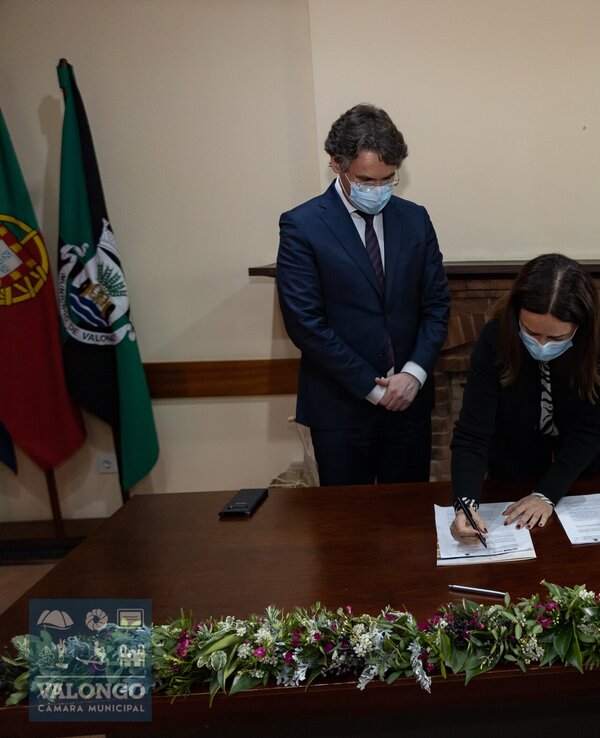 Uma mulher assina documentos em uma cerimónia, com dois homens ao fundo.