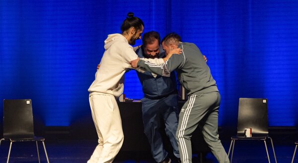 Três pessoas em cena, interagindo em um ambiente teatral iluminado.