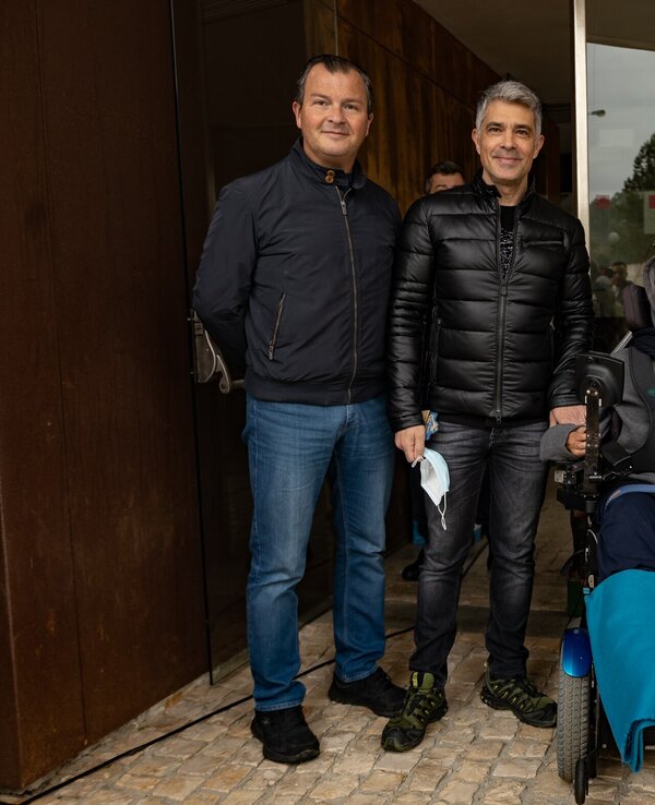 Três pessoas posam em frente a um edifício, uma delas numa cadeira de rodas.