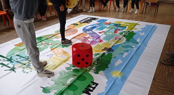 Um jogo de tabuleiro grande no chão, com um dado vermelho ao centro.