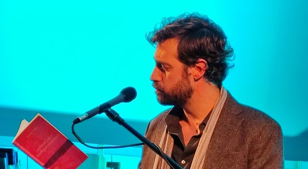 Homem a ler um livro ao microfone, com fundo azul e prateleiras de livros.