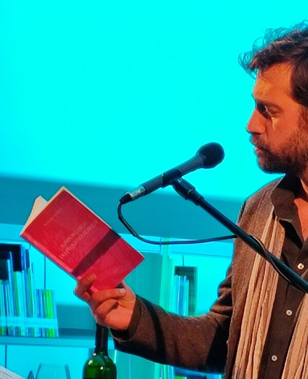 Homem a ler um livro ao microfone, com fundo azul e prateleiras de livros.