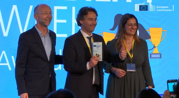 Três pessoas em palco, uma segurando um prémio, com troféus ao fundo.