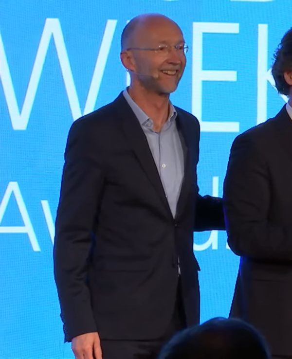 Três pessoas em palco, uma segurando um prémio, com troféus ao fundo.