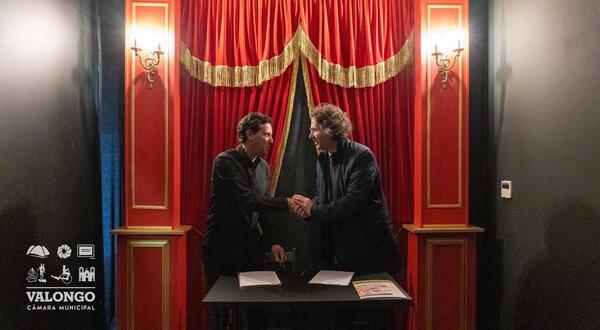 Duas pessoas a apertar as mãos em frente a um fundo de cortinas vermelhas.