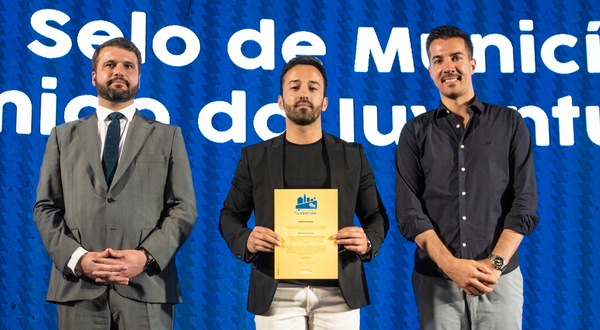 Três homens em palco, um deles segurando um certificado em destaque.