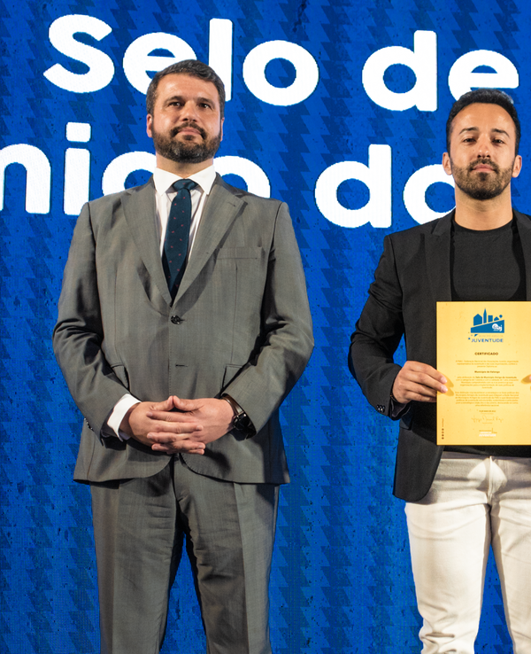 Três homens em palco, um deles segurando um certificado em destaque.