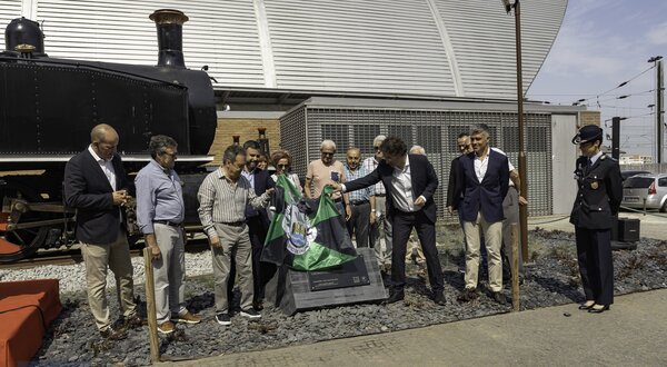 Grupo de pessoas a descobrir uma placa em frente a uma locomotiva histórica.