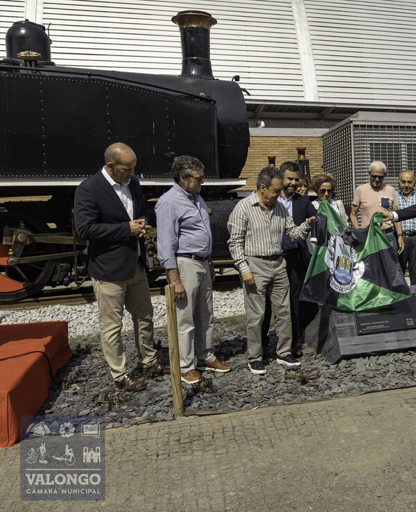 Grupo de pessoas a descobrir uma placa em frente a uma locomotiva histórica.