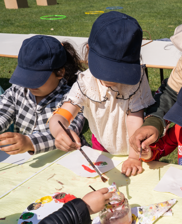 Crianças a pintar com a ajuda de um adulto, em ambiente ao ar livre.